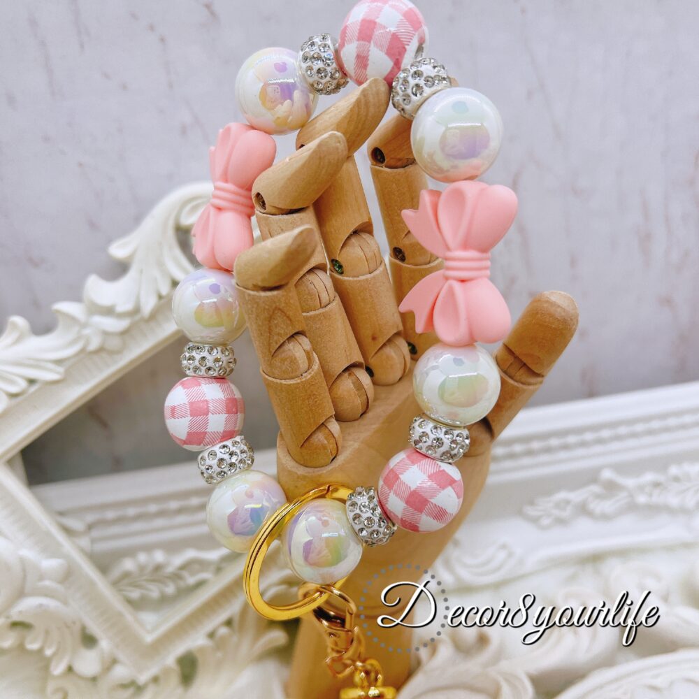 Elegant wristlet keychain featuring pink and white checkered acrylic beads, rhinestone spacers, and a faux-leather tassel displayed on a decorative hand model against a soft, luxurious background.