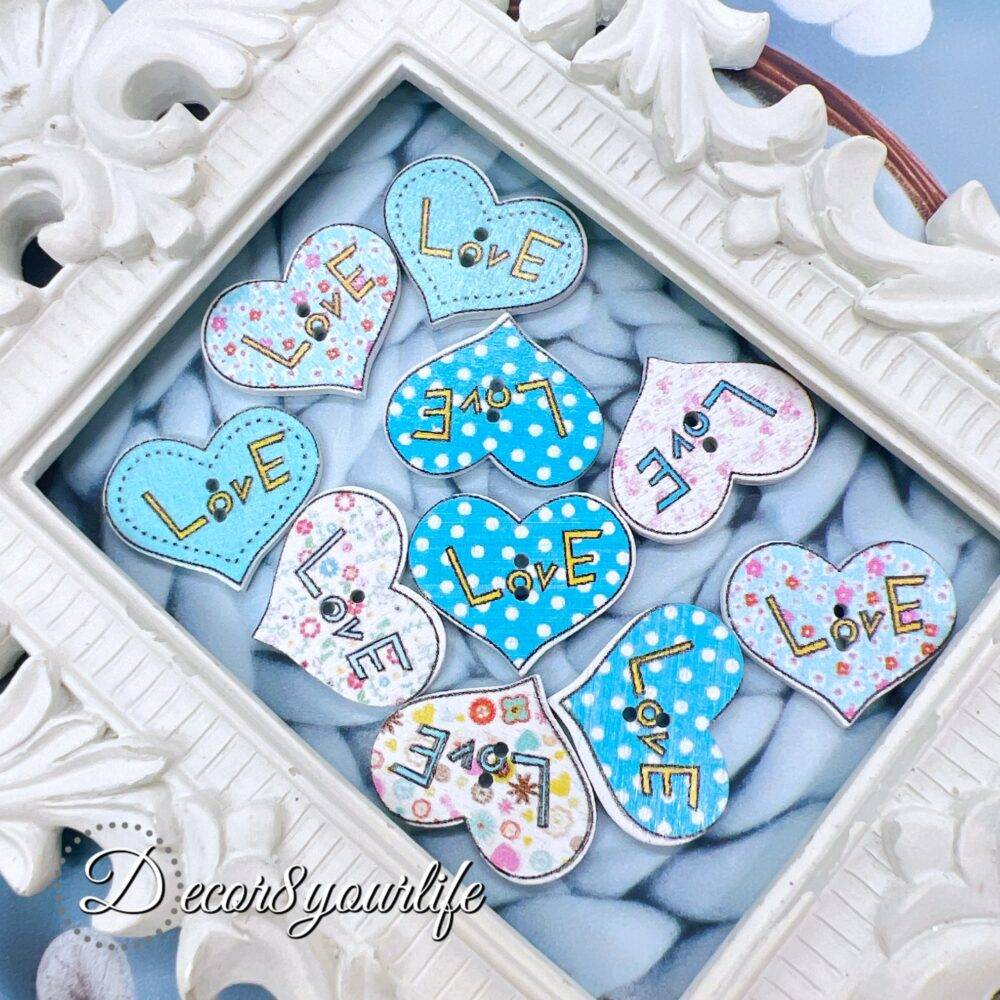 A set of ten heart-shaped wooden buttons with a pink "LOVE" design, displayed inside a white ornate frame. These 1-inch buttons are perfect for card-making, scrapbooking, journaling, and mixed media projects.