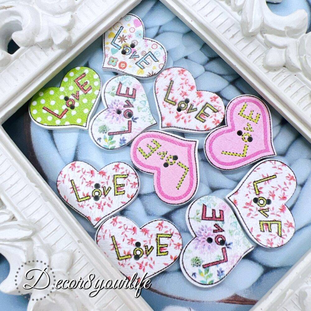 A set of ten heart-shaped wooden buttons with a pink "LOVE" design, displayed inside a white ornate frame. These 1-inch buttons are perfect for card-making, scrapbooking, journaling, and mixed media projects.