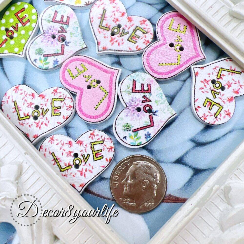 A set of ten heart-shaped wooden buttons with a pink "LOVE" design, displayed inside a white ornate frame. These 1-inch buttons are perfect for card-making, scrapbooking, journaling, and mixed media projects.