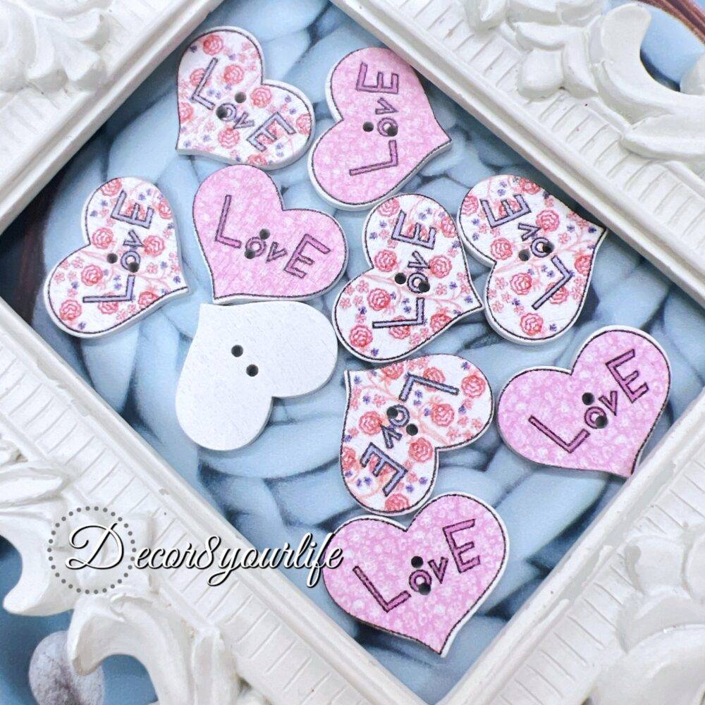 A set of ten heart-shaped wooden buttons with a pink "LOVE" design, displayed inside a white ornate frame. These 1-inch buttons are perfect for card-making, scrapbooking, journaling, and mixed media projects.