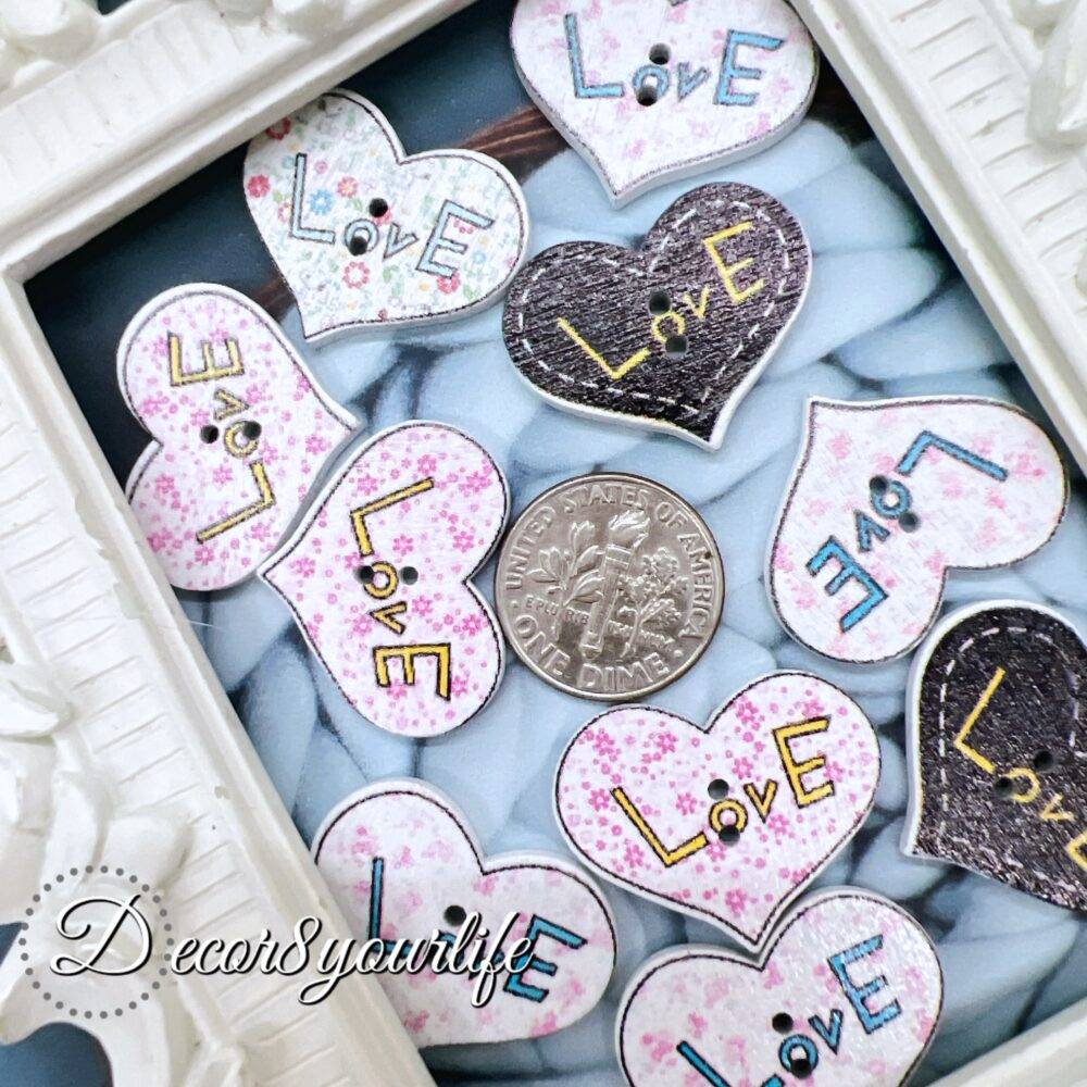 A set of ten heart-shaped wooden buttons with a pink "LOVE" design, displayed inside a white ornate frame. These 1-inch buttons are perfect for card-making, scrapbooking, journaling, and mixed media projects.