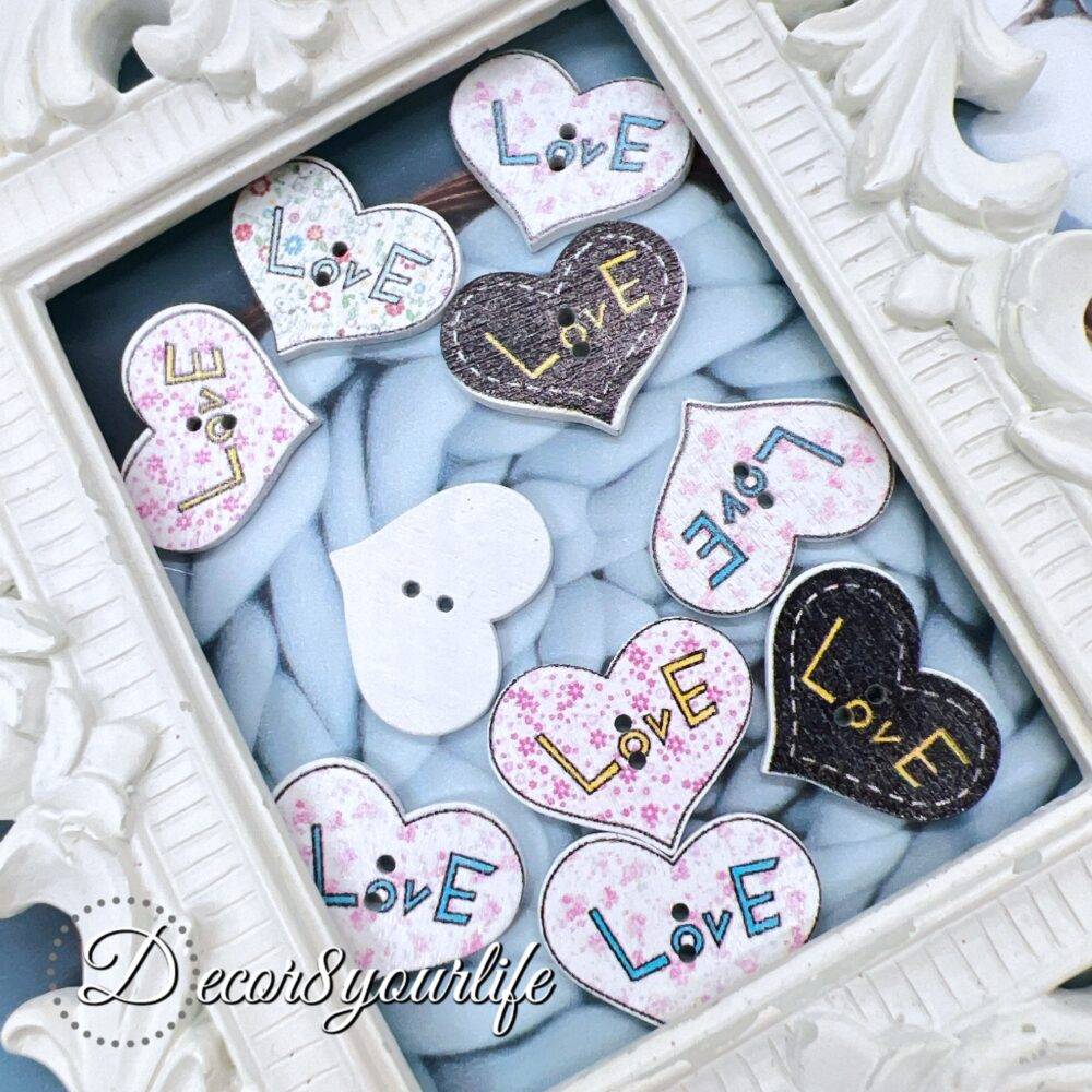 A set of ten heart-shaped wooden buttons with a pink "LOVE" design, displayed inside a white ornate frame. These 1-inch buttons are perfect for card-making, scrapbooking, journaling, and mixed media projects.