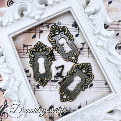 ornate vintage-style keyhole cover with an antique bronze finish, displayed on a decorative sheet of aged musical notes. The keyhole cover features intricate detailing, evoking an old-world charm, and is accompanied by small matching screws. A U.S. quarter coin is placed beside the keyhole cover for size reference. The background includes a white ornate picture frame for an elegant touch.