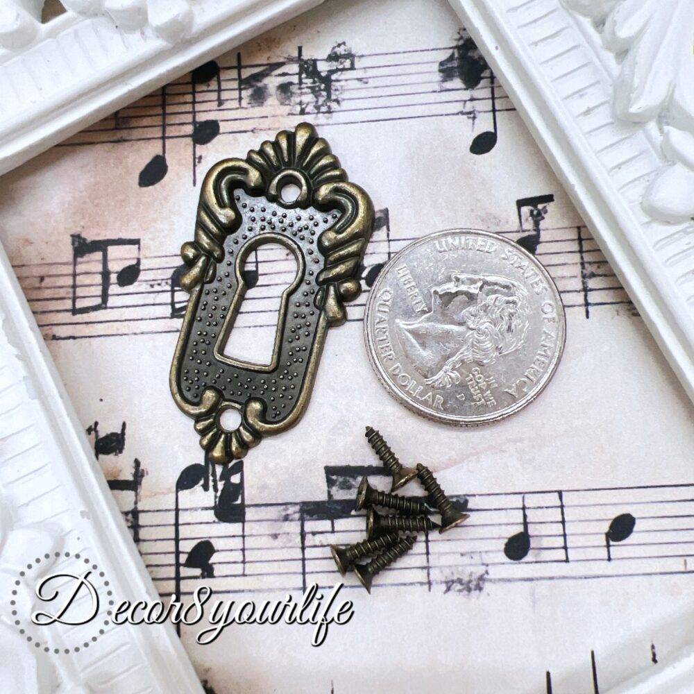ornate vintage-style keyhole cover with an antique bronze finish, displayed on a decorative sheet of aged musical notes. The keyhole cover features intricate detailing, evoking an old-world charm, and is accompanied by small matching screws. A U.S. quarter coin is placed beside the keyhole cover for size reference. The background includes a white ornate picture frame for an elegant touch.