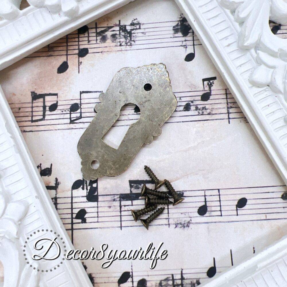 ornate vintage-style keyhole cover with an antique bronze finish, displayed on a decorative sheet of aged musical notes. The keyhole cover features intricate detailing, evoking an old-world charm, and is accompanied by small matching screws. A U.S. quarter coin is placed beside the keyhole cover for size reference. The background includes a white ornate picture frame for an elegant touch.