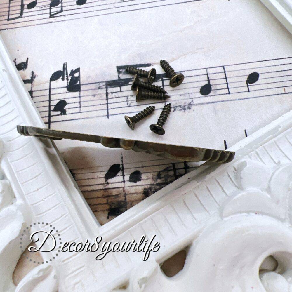 ornate vintage-style keyhole cover with an antique bronze finish, displayed on a decorative sheet of aged musical notes. The keyhole cover features intricate detailing, evoking an old-world charm, and is accompanied by small matching screws. A U.S. quarter coin is placed beside the keyhole cover for size reference. The background includes a white ornate picture frame for an elegant touch.