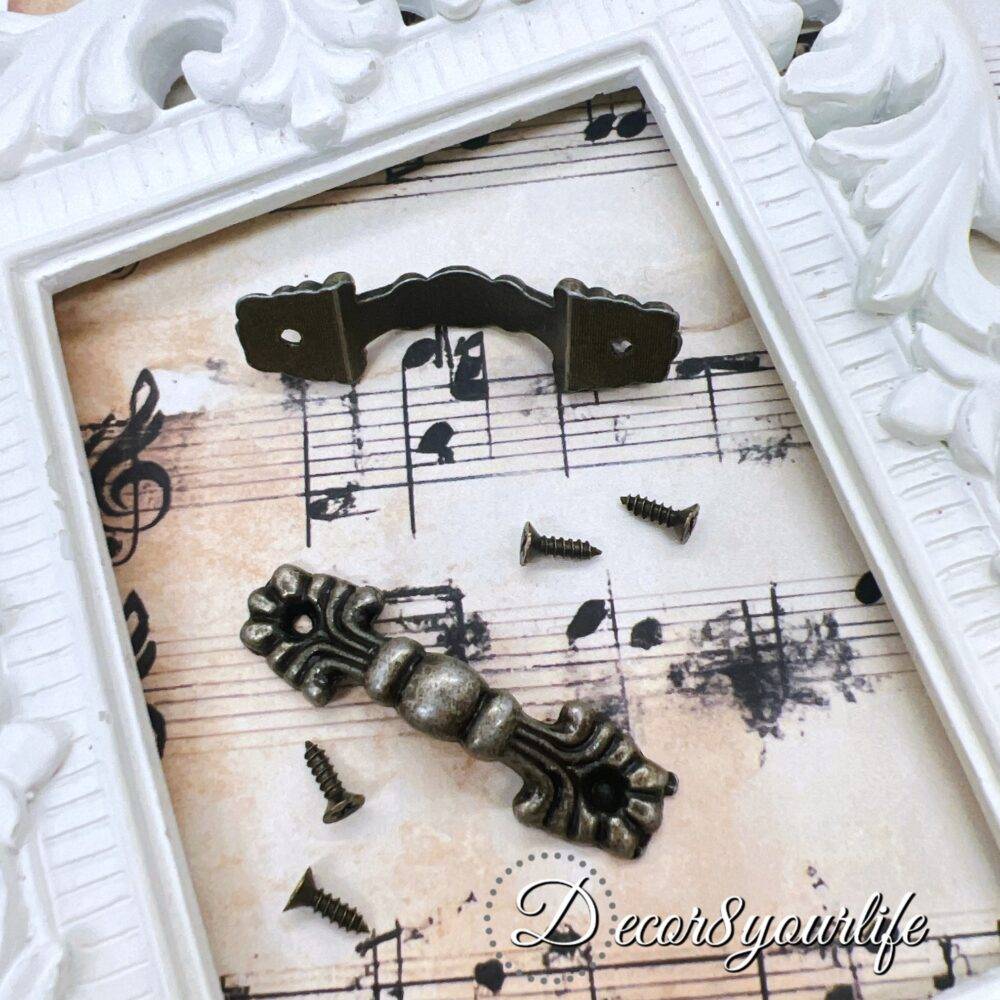 set of two vintage-style drawer handles with an antique bronze finish, displayed on a decorative sheet of music paper within a white ornate frame. Small matching screws are included for easy attachment, making them perfect for DIY furniture restoration, mini album projects, or decorative crafts.