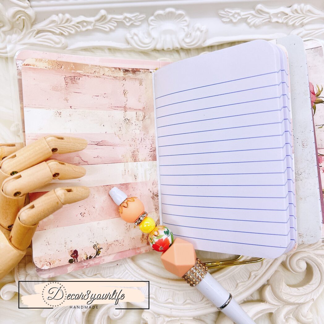 Interior of vintage-style laminated traveler's notebook showing two embellished mini composition books, floral and stripe designs, and a pastel beaded pen on clip