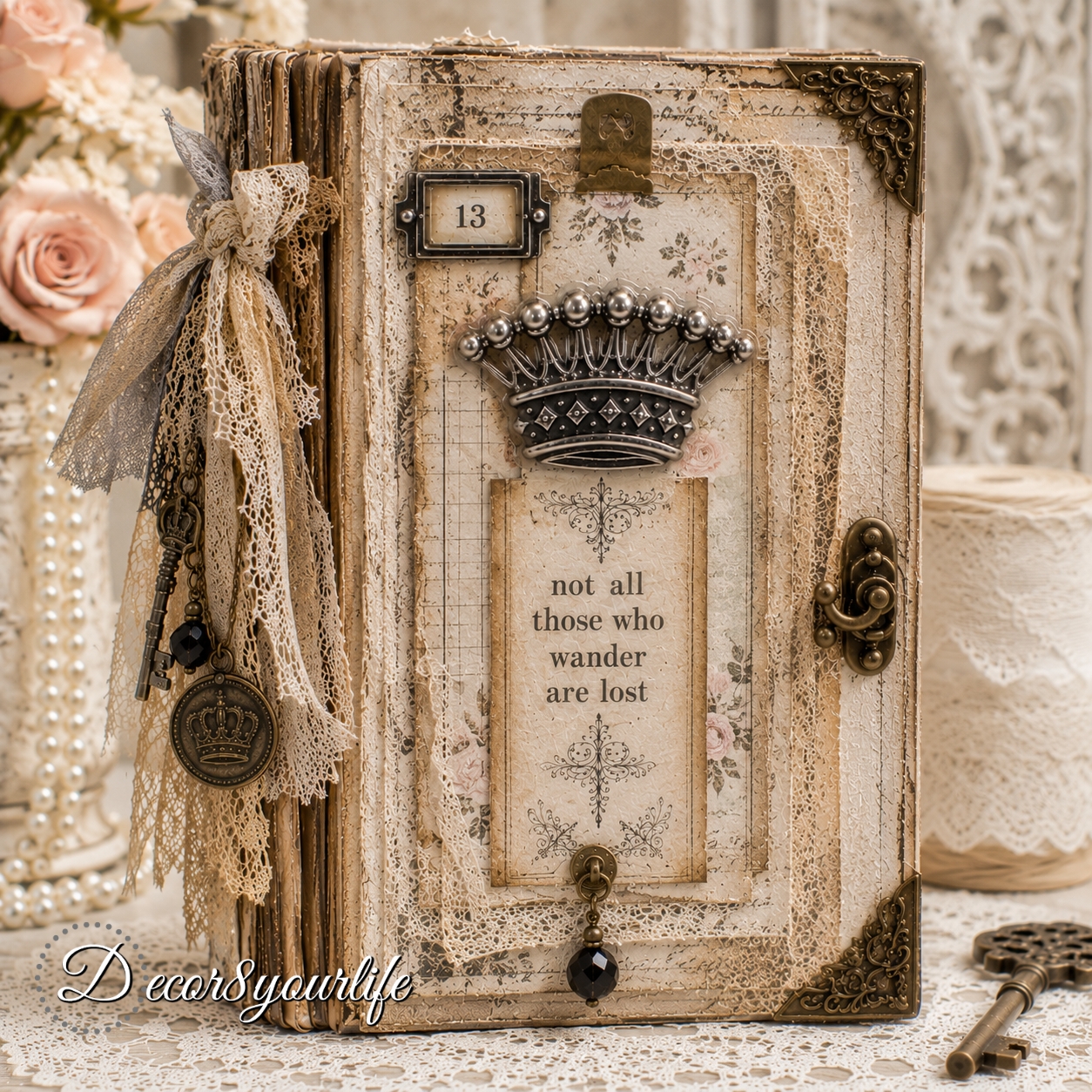 Elegant handmade junk journal with a vintage Victorian-style cover featuring layered cream and floral papers, delicate lace, an antique bronze crown embellishment, ornate metal corner pieces, a “not all those who wander are lost” sentiment panel, and decorative charms against a soft cream shabby chic craft background.